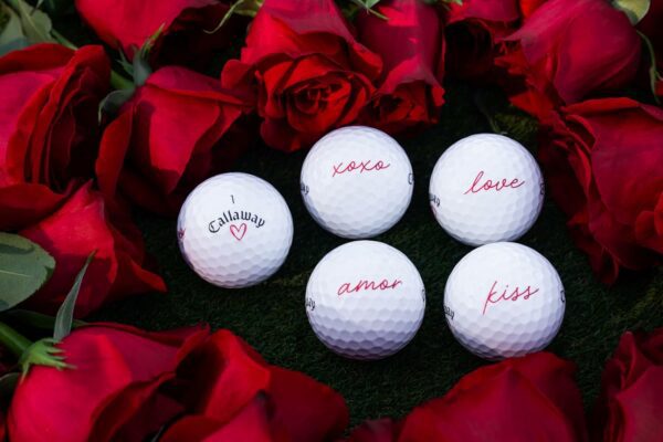 Five white Callaway golf balls on green grass surrounded by red roses, featuring romantic words like xoxo, love, amor, and kiss