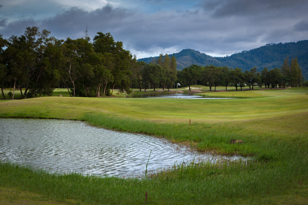 Wide view across water and fairways at Laguna Golf Phuket with hills behind under a stormy sky